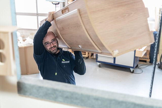 Tischer Manufaktur - Mitarbeiter hebt gebogenes Möbelstück an – Innenausbau in der Werkstatt.