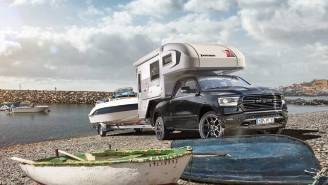 Pick-up mit Tischer-Wohnkabine am Wasser, zieht Motorboot auf Trailer – Küstenlandschaft im Hintergrund.