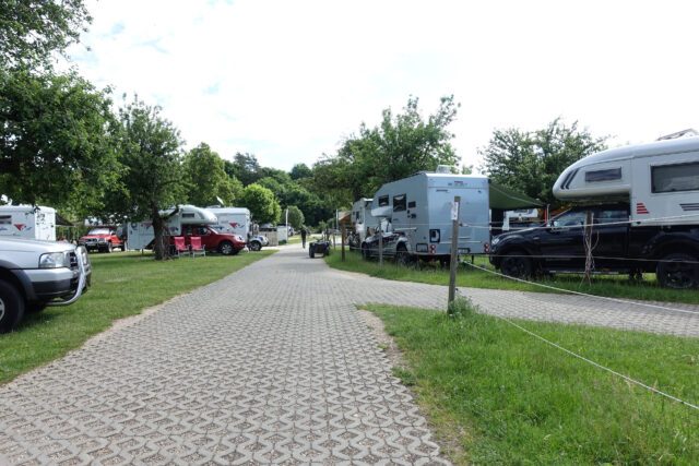 Blick entlang eines Stellplatzes mit aufgereihten Tischer-Kabinen unter Bäumen – sommerliche Stimmung.
