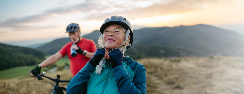 Portrait of beautiful senior couple on bike ride in autumn nature. Taking break, drinking water from sport bottle and fastening helmet.